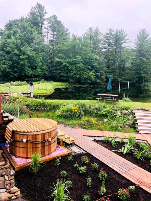 A serene outdoor scene featuring a wooden hot tub, lush greenery, and a tranquil pond surrounded by trees.