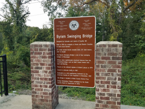 Sign for Byram Swinging Bridge, detailing its history and design, surrounded by trees and greenery.