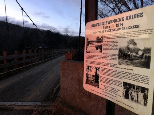 Historic swinging bridge built in 1914, spanning Sylamore Creek, with an informational sign about its history.