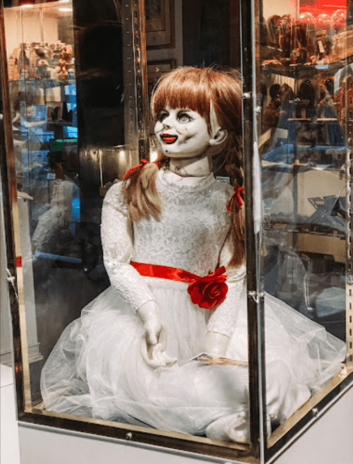 A creepy doll in a glass display case, wearing a white dress with a red ribbon and a rose, with a sinister smile.