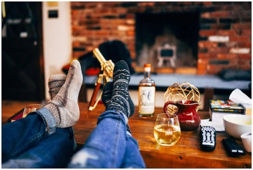 Cozy scene with two pairs of feet in socks, a drink, and a warm fireplace in the background.