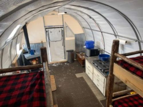 Interior of a cozy cabin with bunk beds, a kitchen area, and a wood stove, featuring rustic decor and a warm atmosphere.