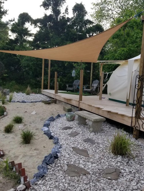 A wooden deck with a tan shade sail, surrounded by gravel and plants, leading to a tent in a green outdoor setting.