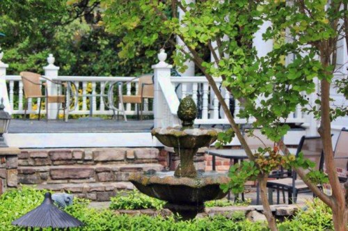 A stone fountain surrounded by greenery, with a patio and chairs visible in the background.