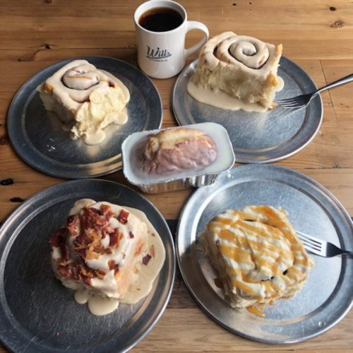 A variety of cinnamon rolls on metal plates, topped with icing and bacon, alongside a cup of coffee.