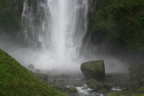 A powerful waterfall cascades down rocky cliffs, surrounded by lush greenery and mist rising from the water.
