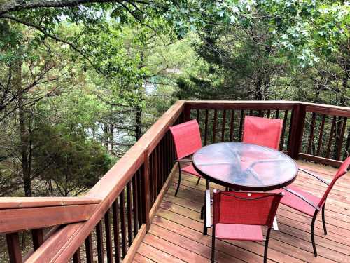 A wooden deck with a round table and four red chairs, surrounded by trees and greenery.