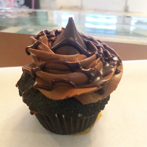 A chocolate cupcake with rich chocolate frosting and a glossy chocolate drizzle on top, sitting on a white surface.