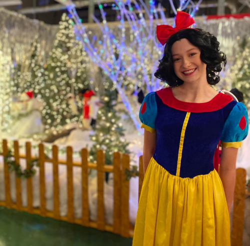 A person dressed as Snow White stands in front of a festive winter scene with lights and decorations.