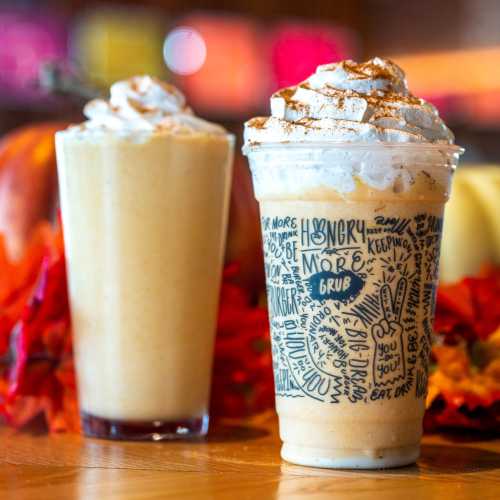 Two festive drinks topped with whipped cream and spices, surrounded by autumn leaves and warm colors.