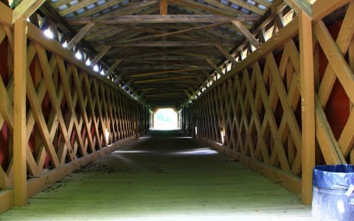 A view inside a covered bridge, featuring wooden beams and a lighted pathway leading to an opening at the end.