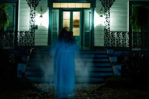 A ghostly figure in a white dress stands on the steps of a dimly lit house, surrounded by shadows and eerie lighting.
