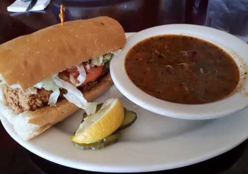 A sandwich with fried chicken, lettuce, and tomato, served with a bowl of soup and a side of pickles and lemon.