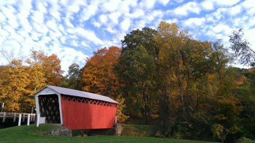 A red covered bridge surrounded by vibrant autumn trees under a cloudy blue sky.
