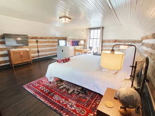 Cozy bedroom with a log cabin design, featuring a bed, TV, and rustic decor, illuminated by natural light.
