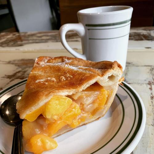 A slice of peach pie on a plate next to a white cup with a green stripe, set on a rustic table.