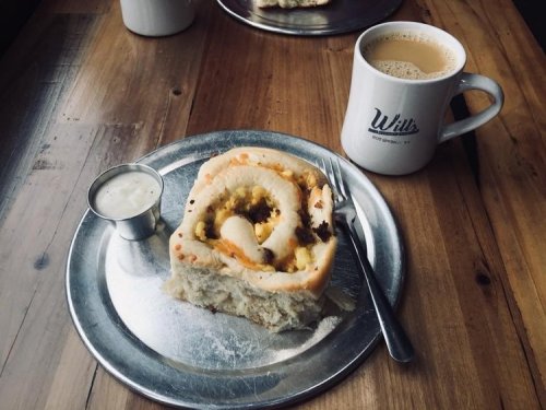 A cinnamon roll on a metal plate with a small cup of cream and a coffee mug on a wooden table.