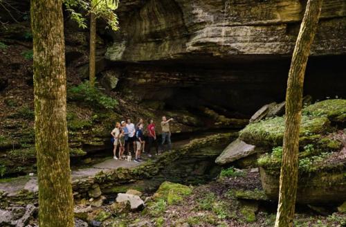 A group of five people stands near a rocky cave in a lush forest, surrounded by trees and greenery.