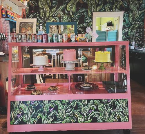 A colorful bakery display case with cakes and pastries, set against a vibrant leaf-patterned wall.