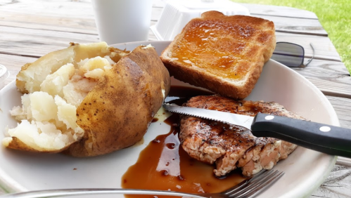 A plate with a baked potato, grilled salmon, and toast with jelly, set on a wooden table outdoors.