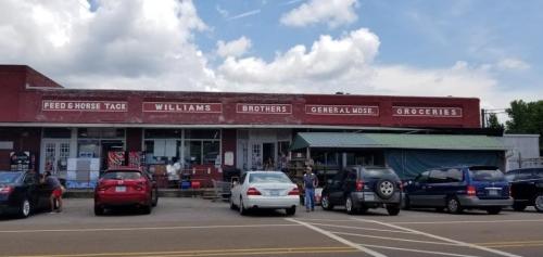 A vintage storefront with signs for "Williams Brothers" and "General Merchandise," surrounded by parked cars.