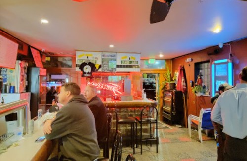 A cozy bar interior with patrons seated at the counter, neon lights, and colorful decor.