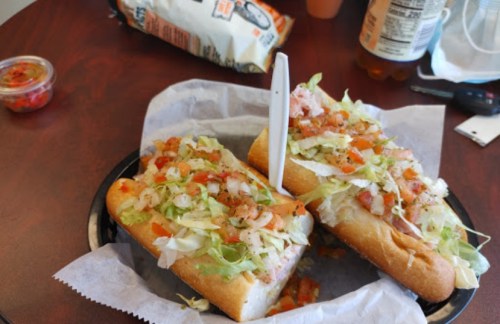 Three loaded sandwiches with lettuce, tomatoes, and toppings on a plate, with a side of salsa and snacks in the background.