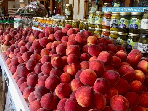 A vibrant display of fresh peaches stacked high on a market table, with jars of pickles in the background.