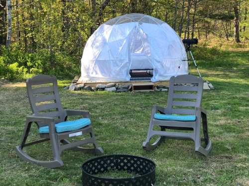 A geodesic dome in a grassy area, with two rocking chairs and a fire pit in front, surrounded by trees.