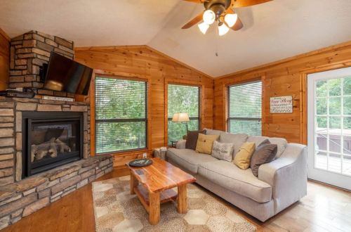 Cozy living room with wooden walls, a stone fireplace, a gray sofa, and large windows letting in natural light.