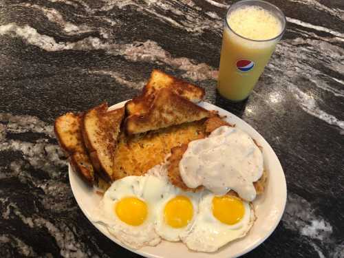 A plate of fried eggs, toast, and a biscuit with gravy, alongside a glass of orange soda.