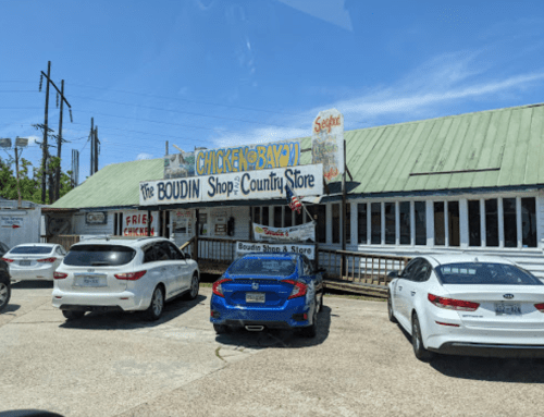 A country store with a green roof, featuring signs for "Chicken Bayou" and "Boudin Shop," surrounded by parked cars.