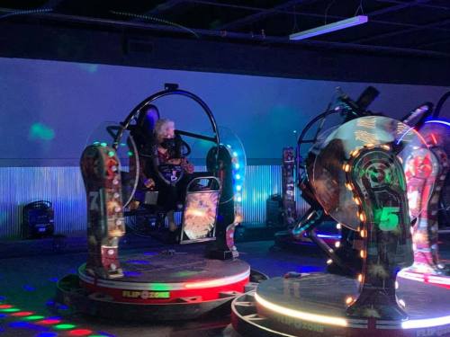 A person sits in a colorful, illuminated arcade ride with spinning seats and vibrant lights in a dark room.