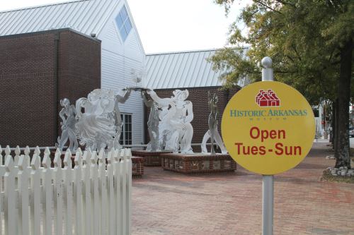 Sign for the Historic Arkansas Museum with a sculpture display in the background and a white picket fence.