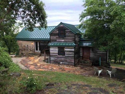 A rustic two-story house with a green metal roof, surrounded by trees and a stone pathway.