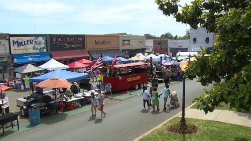 A bustling street market with colorful tents, food vendors, and people walking, set in a small town.
