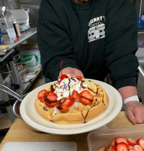 A waffle topped with whipped cream, strawberries, and sprinkles, served with a side of fruit in a diner setting.