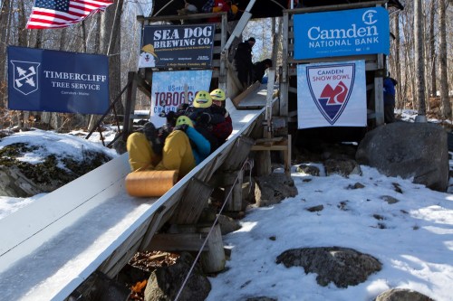 Two people slide down a snowy ramp on a tube, with banners for local businesses in the background.