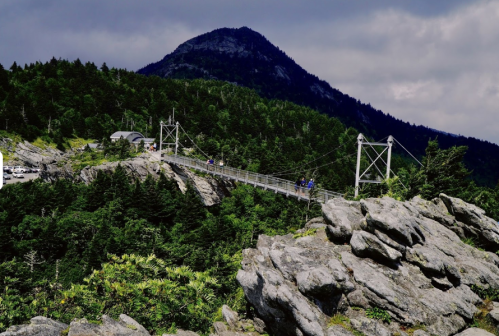 A suspension bridge spans rocky terrain, surrounded by lush green trees and a mountain in the background under a cloudy sky.