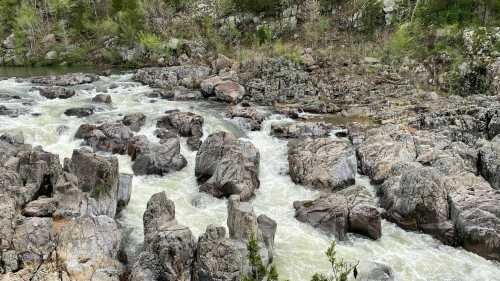 A rushing river flows over rocky terrain, surrounded by greenery and rugged cliffs.