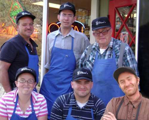 A group of six people in blue aprons and hats, smiling outside a store, with a welcoming atmosphere.