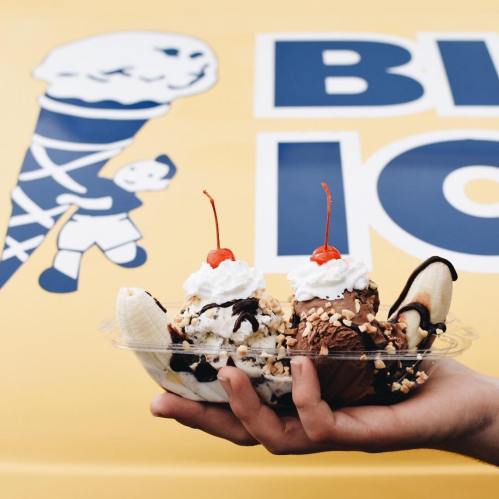A hand holding a banana split topped with whipped cream, nuts, and cherries, in front of an ice cream truck.