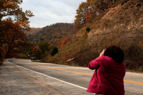 A person takes a photo of a scenic road surrounded by autumn foliage and hills in the background.