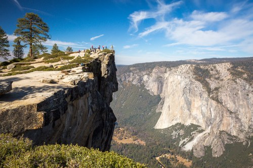 A scenic view from a cliff edge overlooking a vast valley and mountains under a blue sky with wispy clouds.