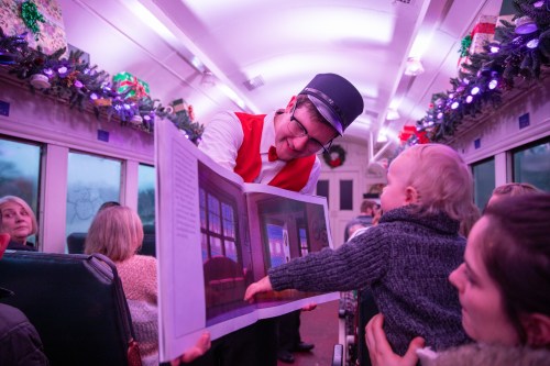 A conductor shows a storybook to a child on a festive train, surrounded by holiday decorations and smiling passengers.