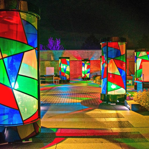 Colorful illuminated pillars with stained glass patterns in a nighttime outdoor setting, casting vibrant reflections on the ground.