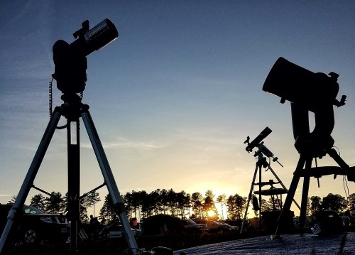 Silhouettes of telescopes against a sunset sky, with trees in the background and a clear horizon.