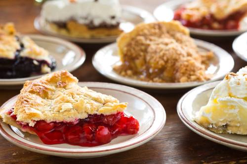 A variety of pie slices on plates, including cherry, chocolate, and apple, arranged on a wooden table.
