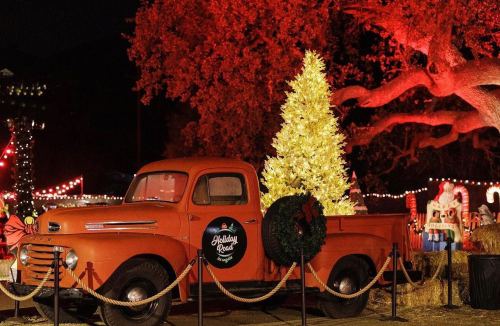 An orange vintage truck with a yellow Christmas tree in the bed, surrounded by festive decorations and lights.