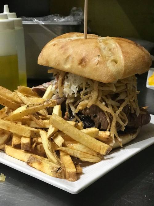 A large sandwich piled with meat and crispy toppings, served with a side of golden French fries on a white plate.